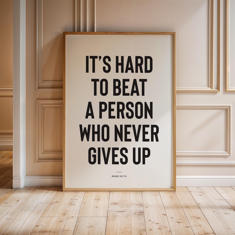 Puede incluir: Un cartel enmarcado con la cita "IT'S HARD TO BEAT A PERSON WHO NEVER GIVES UP" en letras negras en negrita. La cita se atribuye a Babe Ruth. El marco es de color madera clara.