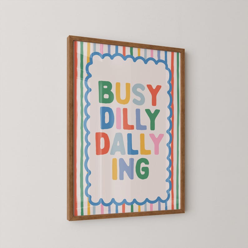 May include: A framed art print with the words "BUSY DILLY DALLY ING" in colorful block letters. The artwork has a white background with a scalloped border and is framed in a brown wooden frame. The background has vertical stripes.