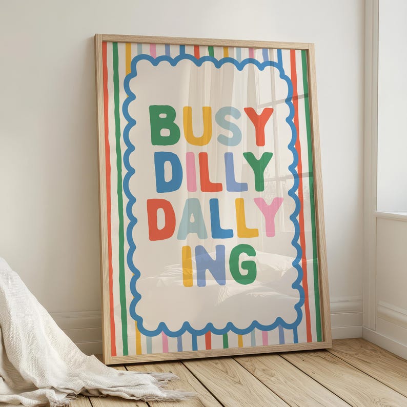 May include: A framed art print with the words "BUSY DILLY DALLY ING" in colorful block letters. The print has a cream background with a blue scalloped border and colorful vertical stripes. The frame is made of light-colored wood.