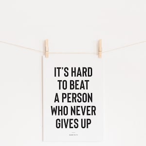 Puede incluir: Un p&oacute;ster blanco con la cita "IT'S HARD TO BEAT A PERSON WHO NEVER GIVES UP" en letras negras en negrita, atribuida a Babe Ruth. El p&oacute;ster est&aacute; suspendido por dos pinzas de madera en una cuerda fina sobre un fondo blanco.