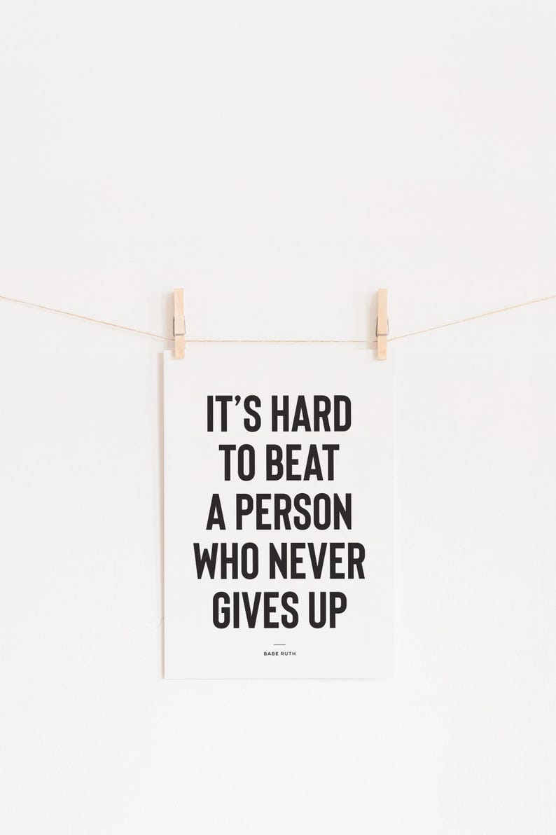 Puede incluir: Un p&oacute;ster blanco con la cita "IT'S HARD TO BEAT A PERSON WHO NEVER GIVES UP" en letras negras en negrita, atribuida a Babe Ruth. El p&oacute;ster est&aacute; suspendido por dos pinzas de madera en una cuerda fina sobre un fondo blanco.