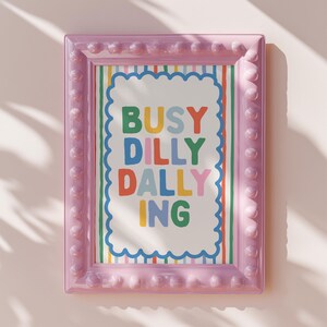 May include: A framed art print with the words "BUSY DILLY DALLY ING" in colorful letters. The print is in a pink frame with a bubble-like border, and the background has a striped pattern.