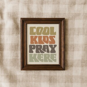 May include: A framed print with the words "COOL KIDS PRAY HERE" in a retro font. The text is in shades of green, brown, and orange against a cream background. The frame is dark brown.