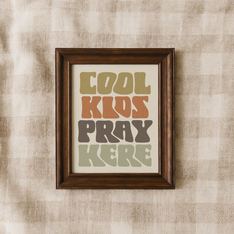 May include: A framed print with the words "COOL KIDS PRAY HERE" in a retro font. The text is in shades of green, brown, and orange against a cream background. The frame is dark brown.