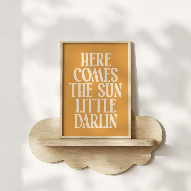 K&ouml;nnte beinhalten: Ein gerahmter Druck mit gelbem Hintergrund und wei&szlig;em Text, der "Here comes the sun little darlin" lautet. Der Druck befindet sich auf einem wolkenf&ouml;rmigen Holzregal.