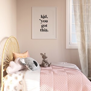 May include: A pink bed with a white and yellow rainbow patterned sheet and a pink quilted blanket. The bed has a wicker headboard and is decorated with pillows and stuffed animals. A framed print with the text "kid, you got this." hangs on the wall behind the bed.