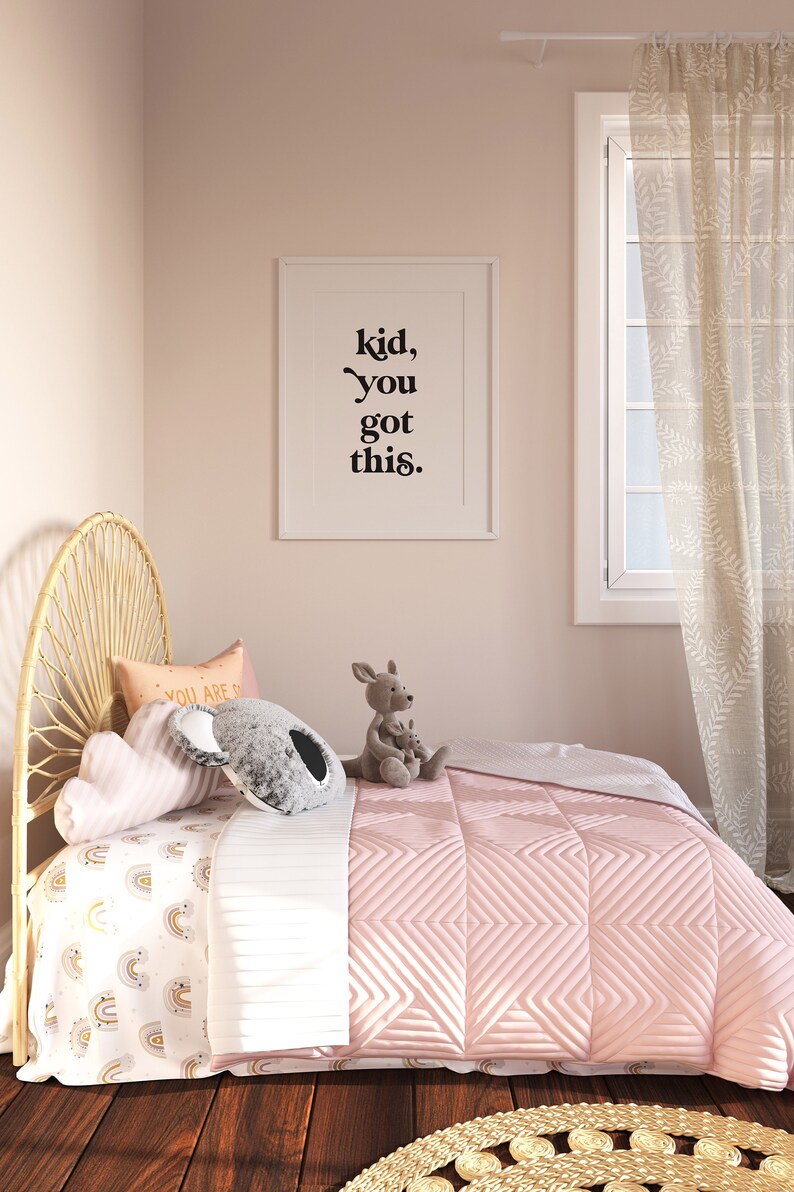 May include: A pink bed with a white and yellow rainbow patterned sheet and a pink quilted blanket. The bed has a wicker headboard and is decorated with pillows and stuffed animals. A framed print with the text "kid, you got this." hangs on the wall behind the bed.