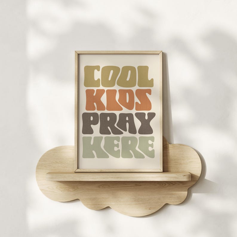 May include: A framed print with the words "COOL KIDS PRAY HERE" in retro-style lettering. The text is in shades of brown, orange, and green. The print is displayed on a cloud-shaped wooden shelf.