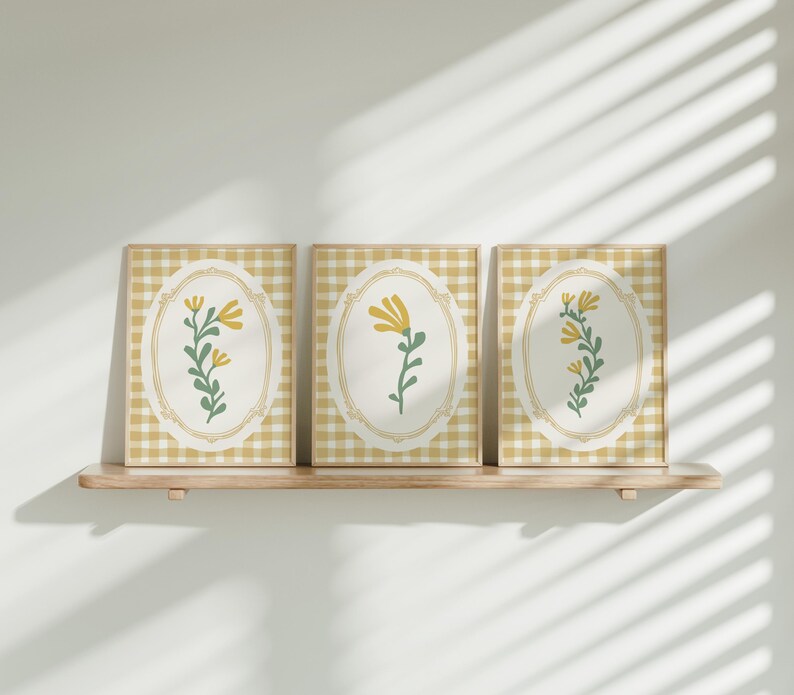 May include: Three framed prints featuring a yellow and green floral design on a white background. The frames are a light wood colour and are displayed on a wooden shelf.