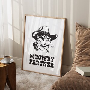 Pu&ograve; includere: Illustrazione in bianco e nero di un gatto con un cappello da cowboy con il testo "MEOWDY PARTNER" sotto. L'illustrazione &egrave; incorniciata in una cornice di legno chiaro.