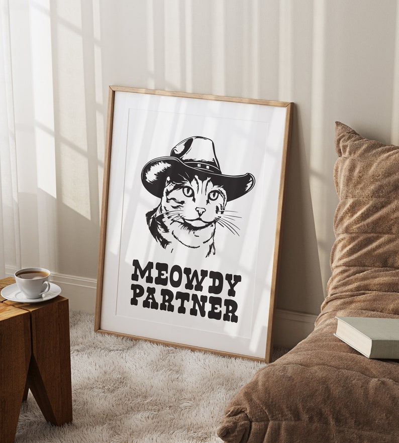 Pu&ograve; includere: Illustrazione in bianco e nero di un gatto con un cappello da cowboy con il testo "MEOWDY PARTNER" sotto. L'illustrazione &egrave; incorniciata in una cornice di legno chiaro.