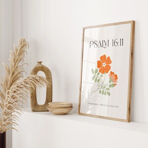 K&ouml;nnte beinhalten: Ein gerahmter Kunstdruck mit einem hellen Holzrahmen. Der Druck zeigt orange und rosa Blumen mit gr&uuml;nen Bl&auml;ttern und den Text "PSALM 16:11" und "IN HIS PRESENCE THERE IS FULLNESS OF JOY". Dekorative Holzschalen und eine Vase sind ebenfalls zu sehen.