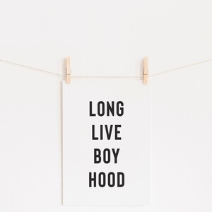 May include: A white poster with black text that reads "LONG LIVE BOY HOOD" hanging on a white wall with two wooden clothespins.