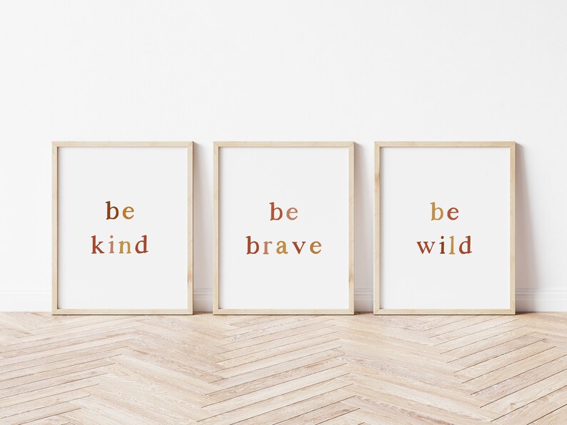May include: Three framed prints with the words "be kind", "be brave", and "be wild" in a gradient of brown and gold colours. The prints are hanging on a white wall with a wood floor.