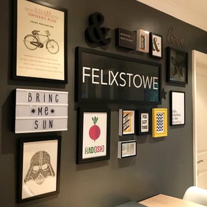 Op de afbeelding: Een zwart-witte muur met verschillende ingelijste prints en kunstwerken. De prints omvatten een fietsveiligheidsplakat, een bord met de tekst "Bring Me Sun", een radijs-illustratie en een bord met de tekst "Felixstowe".