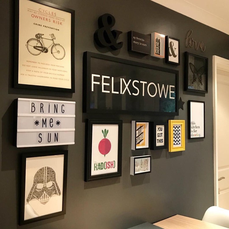Op de afbeelding: Een zwart-witte muur met verschillende ingelijste prints en kunstwerken. De prints omvatten een fietsveiligheidsplakat, een bord met de tekst "Bring Me Sun", een radijs-illustratie en een bord met de tekst "Felixstowe".