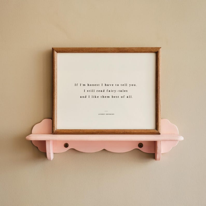 May include: A framed print with a quote about fairy tales rests on a pink, scalloped shelf. The wooden frame surrounds a white background with black text. The shelf is mounted on a neutral-colored wall, creating a simple, elegant display.