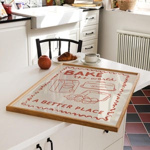 May include: A framed print with a red and white design that says "Bake the world a better place". The print features illustrations of a bag of flour, a baking dish, and a carton of eggs. The frame is made of wood and has a light brown finish.