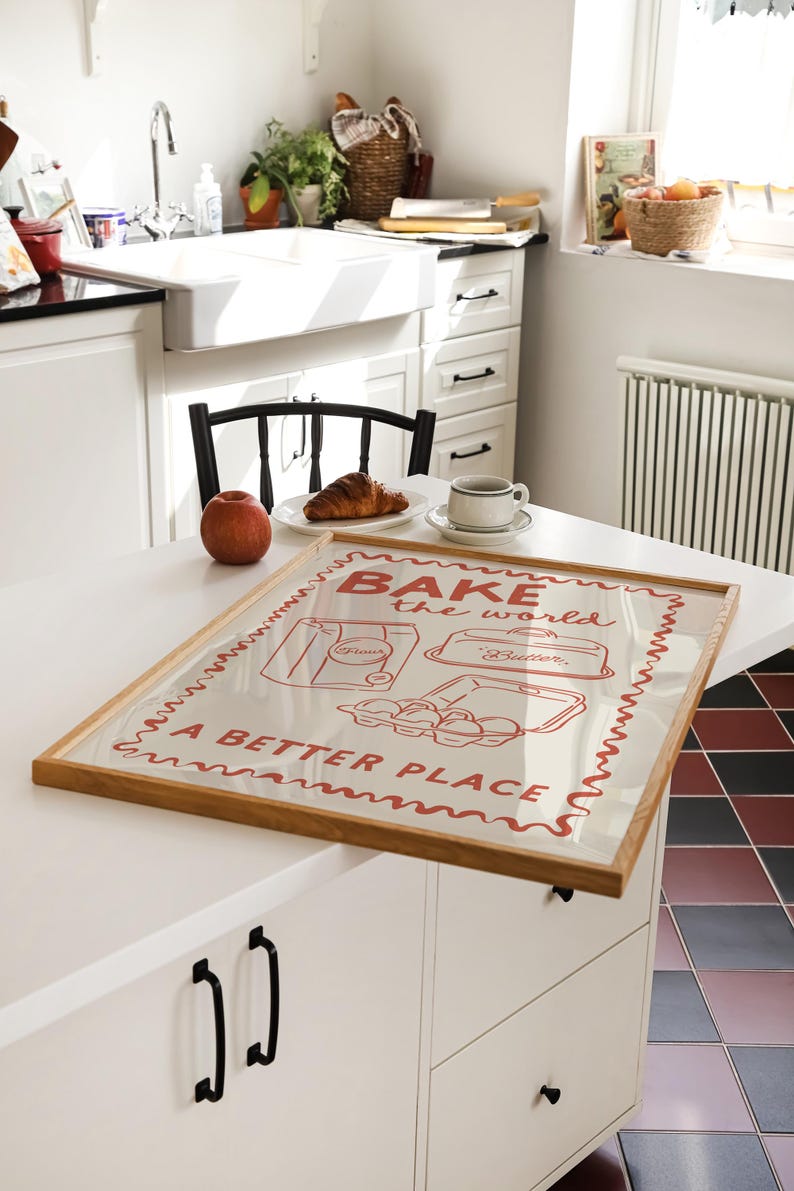May include: A framed print with a red and white design that says "Bake the world a better place". The print features illustrations of a bag of flour, a baking dish, and a carton of eggs. The frame is made of wood and has a light brown finish.