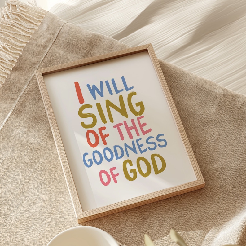 May include: A framed print with the text "I WILL SING OF THE GOODNESS OF GOD" in colorful, handwritten lettering. The frame is light wood and the background is a light beige fabric.