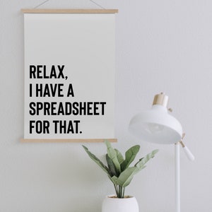 May include: A white poster with black text that reads "Relax, I have a spreadsheet for that." The poster is hanging on a white wall with a small potted plant and a white desk lamp to the right.