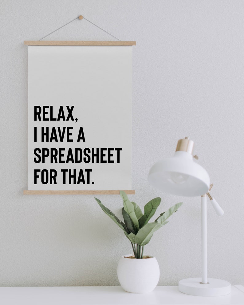 May include: A white poster with black text that reads "Relax, I have a spreadsheet for that." The poster is hanging on a white wall with a small potted plant and a white desk lamp to the right.
