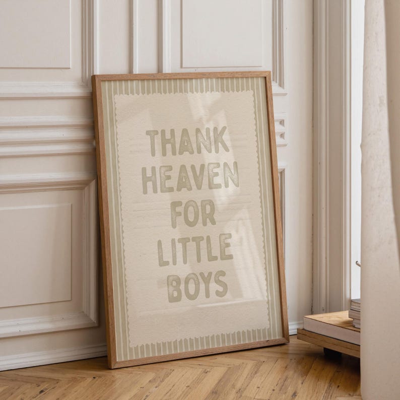 Puede incluir: Un estampado enmarcado con el texto "THANK HEAVEN FOR LITTLE BOYS" en fuente gris claro. El estampado tiene un fondo beige claro con un marco marr&oacute;n claro. El estampado est&aacute; apoyado contra una pared blanca y un suelo de madera.