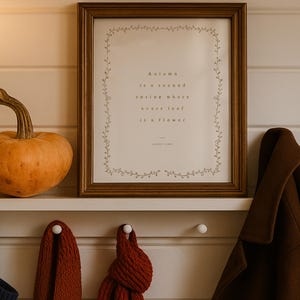 May include: A framed print with the quote "Autumn is a second spring where every leaf is a flower" by Albert Camus. The print is in a brown frame, with a decorative vine border. A pumpkin and a coat are also visible.