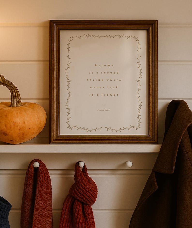 May include: A framed print with the quote "Autumn is a second spring where every leaf is a flower" by Albert Camus. The print is in a brown frame, with a decorative vine border. A pumpkin and a coat are also visible.
