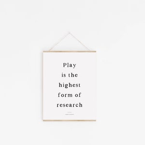 May include: A white framed print with the text "Play is the highest form of research" hanging on a white wall.