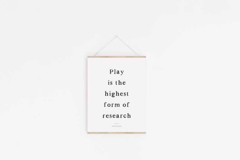 May include: A white framed print with the text "Play is the highest form of research" hanging on a white wall.