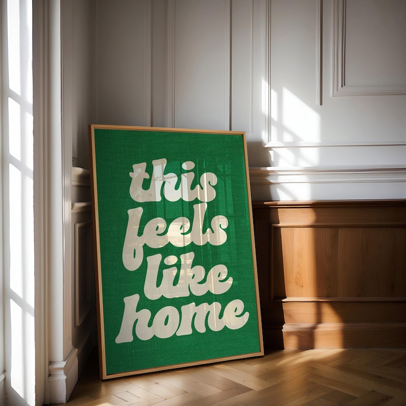 May include: A framed green canvas art print with the words "this feels like home" in a white, retro font. The artwork is displayed on a wooden floor, leaning against a white wall with sunlight streaming in from a window.