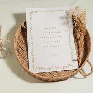 May include: A decorative print with the text "Autumn is a second spring where every leaf is a flower" sits on a woven tray. Dried flowers and a small glass vase with wheat stalks are also present.