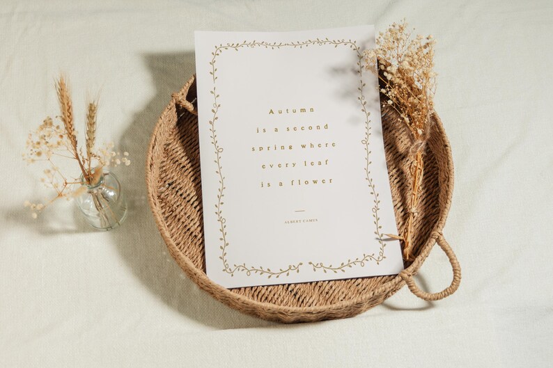 May include: A decorative print with the text "Autumn is a second spring where every leaf is a flower" sits on a woven tray. Dried flowers and a small glass vase with wheat stalks are also present.