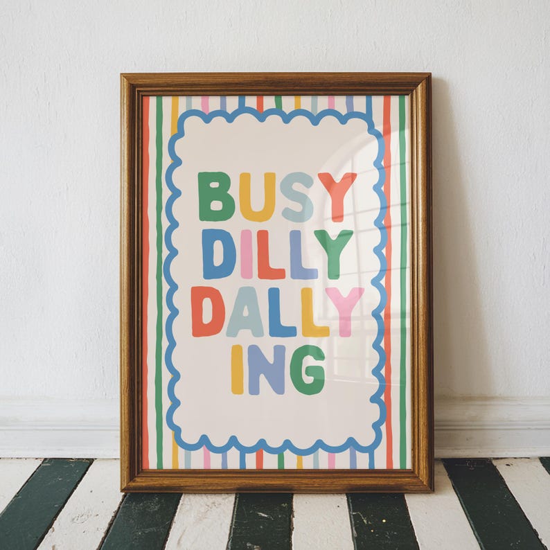 May include: A framed art print with the words "BUSY DILLY DALLY ING" in colorful letters. The text is inside a white scalloped border, set against a striped background of various colors. The frame is a warm brown color.