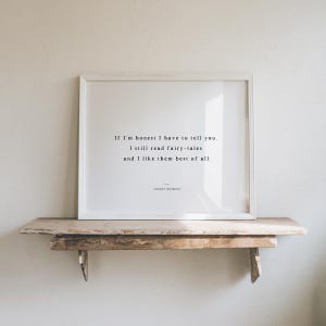 May include: A framed print with a white border displays a quote in black text: "If I'm honest I have to tell you. I still read fairy-tales and I like them best of all." The print rests on a rustic wooden shelf.