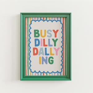May include: A framed art print with the words "BUSY DILLY DALLYING" in colorful block letters. The print has a white background with a scalloped border and is framed in a green frame. The background has vertical stripes.