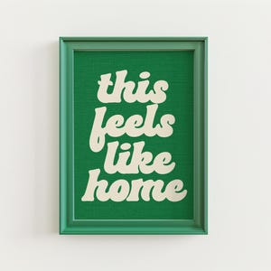 May include: A framed art print with the text "this feels like home" in a retro font. The text is cream-colored against a green background. The frame is green.