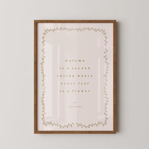 May include: Framed art print with a quote by Albert Camus: "Autumn is a second spring where every leaf is a flower." The print has a light pink background, gold text, and a decorative border. The frame is brown.