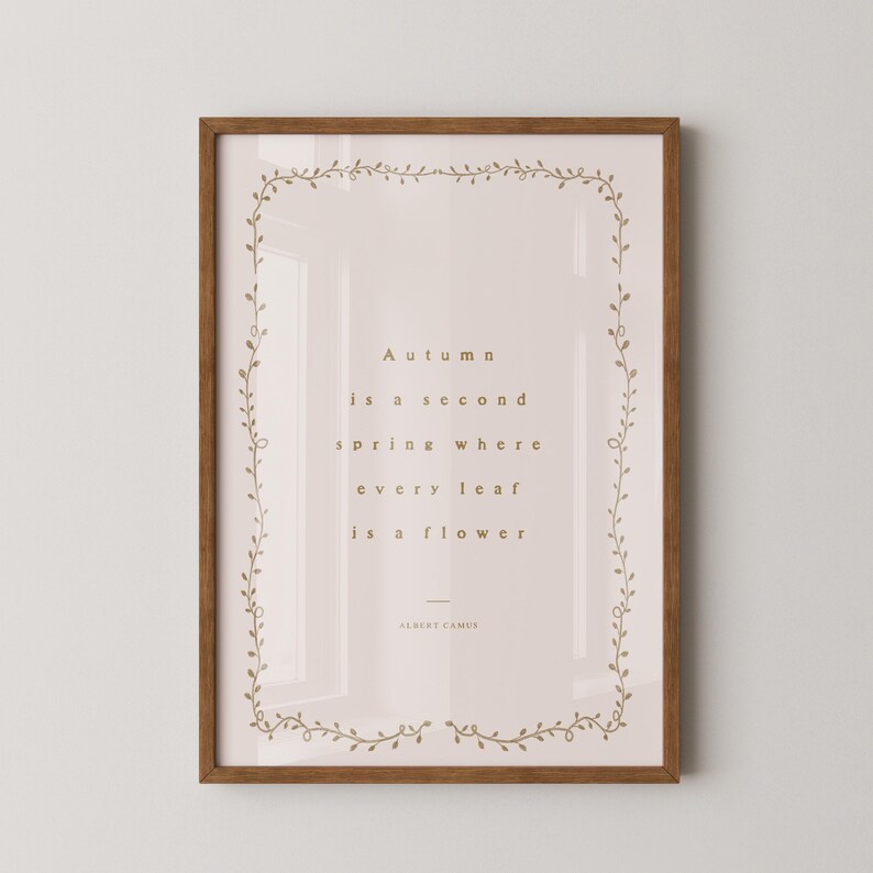 May include: Framed art print with a quote by Albert Camus: "Autumn is a second spring where every leaf is a flower." The print has a light pink background, gold text, and a decorative border. The frame is brown.