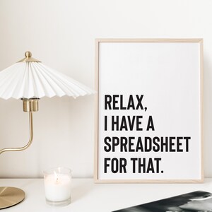 May include: A framed print with the text "Relax, I have a spreadsheet for that." The print is on a white background with a light wood frame. The print is sitting on a white surface with a gold lamp and a white candle.