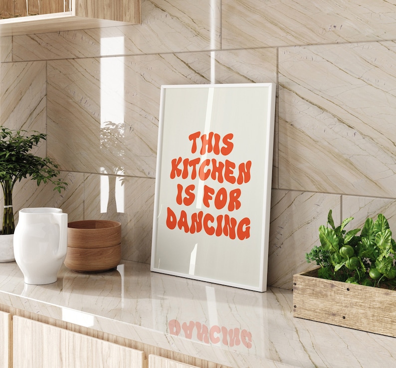 May include: A white framed print with the text "This Kitchen is for Dancing" in red letters on a beige background. The print is hanging on a tiled wall in a kitchen.