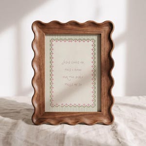 May include: A rectangular wooden frame with a wavy, scalloped edge containing a print. The print features a light beige background with thin vertical stripes and a pink and green floral border, with the text Jesus loves me this I know for the Bible tells me so.