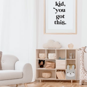 May include: A white framed print with the text "kid, you got this." hanging on a white wall in a nursery. The print is above a wooden shelf with a variety of toys and storage baskets. There is a white armchair in the foreground.