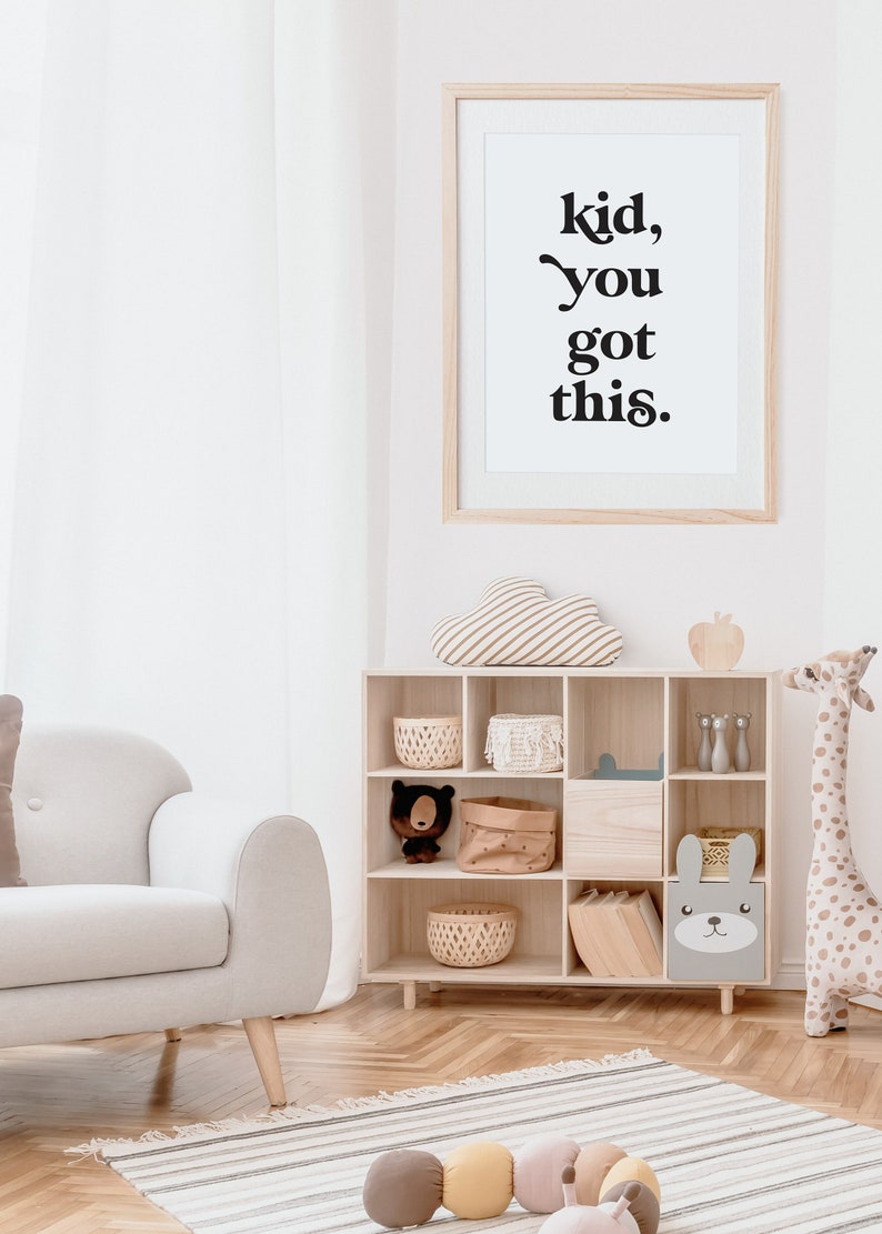 May include: A white framed print with the text "kid, you got this." hanging on a white wall in a nursery. The print is above a wooden shelf with a variety of toys and storage baskets. There is a white armchair in the foreground.