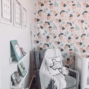 May include: A gray armchair with a white throw blanket and a white pillow with a black outline of the state of Texas and the word "Texas" on it. The armchair is in front of a wall with a lemon-themed wallpaper. There are books on a shelf to the left of the armchair.