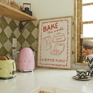 May include: A pink electric kettle and a yellow toaster sit on a white countertop next to a framed print with the text "BAKE the world a BETTER PLACE". The print features illustrations of baking ingredients, including flour, butter, and eggs.