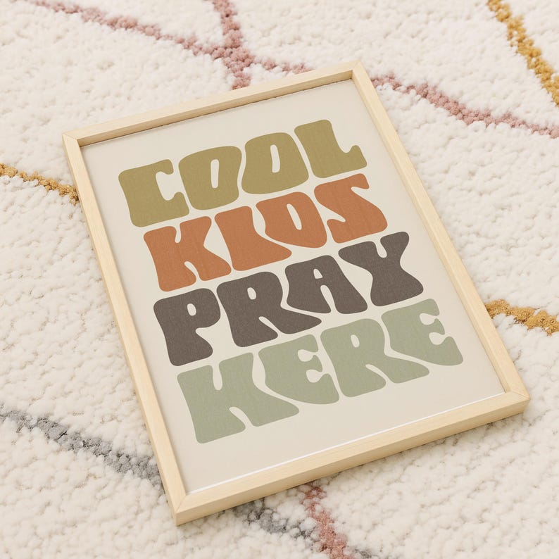 May include: A framed print with the words "COOL KIDS PRAY HERE" in a retro font. The text is in shades of brown, orange, and green on an off-white background. The frame is made of light-colored wood.