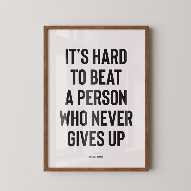 Puede incluir: Un p&oacute;ster enmarcado con la cita "IT'S HARD TO BEAT A PERSON WHO NEVER GIVES UP" en letras negras sobre un fondo blanco. La cita se atribuye a Babe Ruth. El marco es de color marr&oacute;n c&aacute;lido.