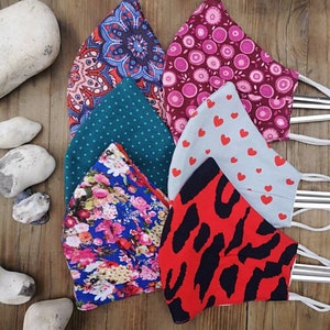 May include: Six different fabric face masks with various patterns. The masks are made of cotton and have a variety of colors, including red, blue, green, pink, and white. The masks are all folded and arranged in a pile on a wooden surface.